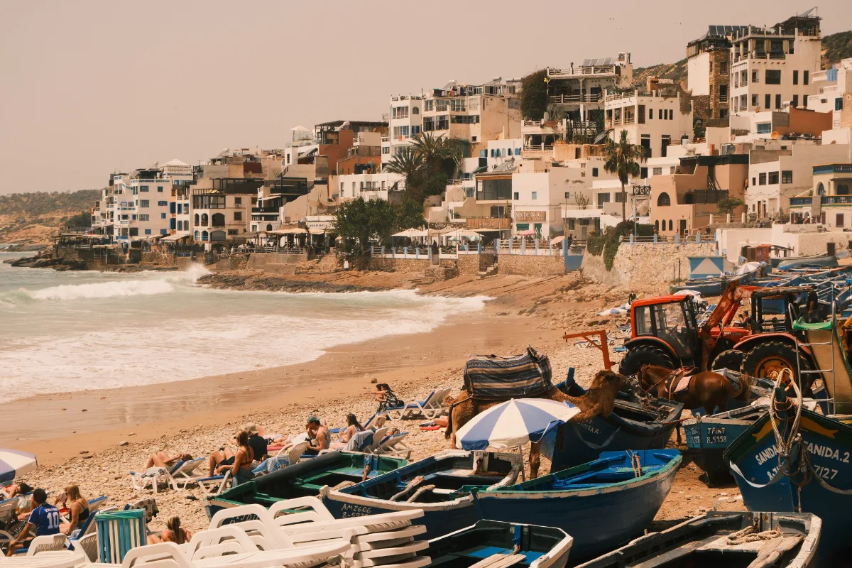 Scenic view of Taghazout beach with colorful boats, sunbathers, and adjacent hillside buildings.